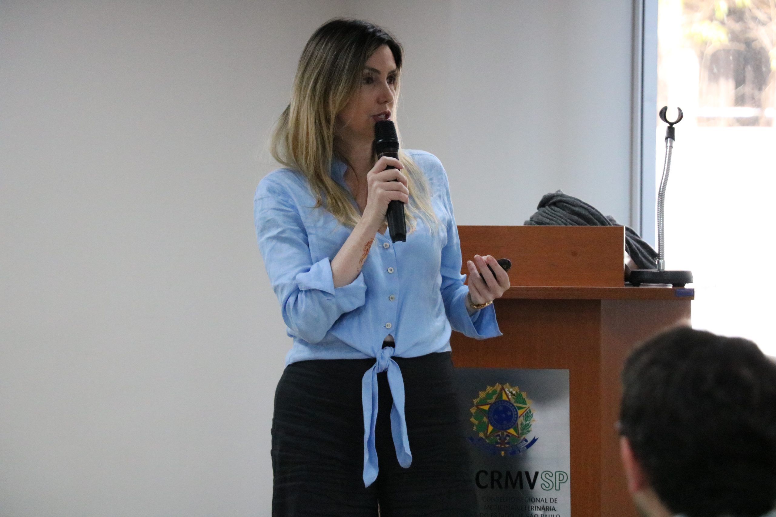 Mulher loira de fios lisos na altura do busto, ela veste blusa azul clara com botões no meio e calça social preta, falando no microfone. É a presidente da Comissão de Atenção à Saúde Mental dos Médicos-veterinários e Zootecnistas, Ingrid Bueno Atayde Machado.