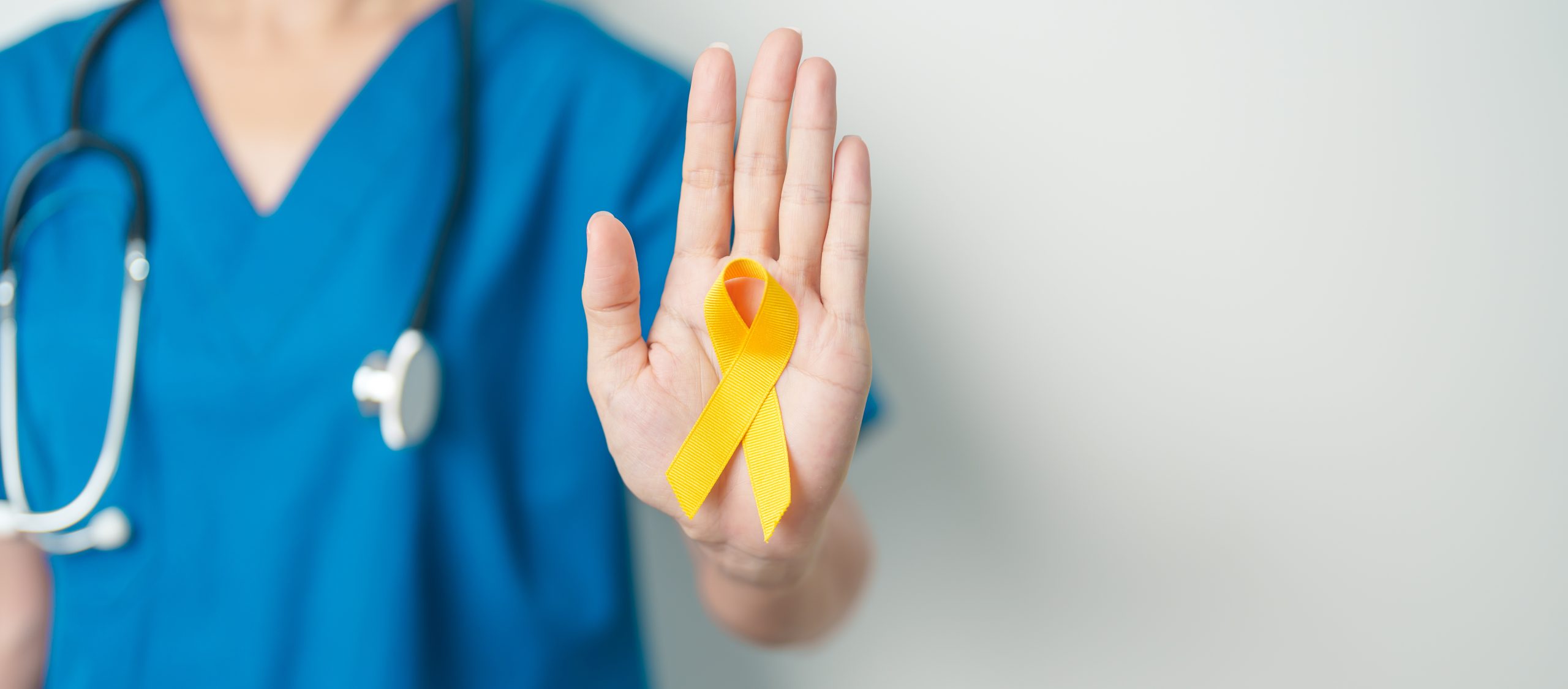 A imagem mostra o corpo de uma médica-veterinária branca, em pé, vestindo uniforme azul e com o estetoscópio no pescoço com o braço direito esticado e a mão direita em riste com um laço amarelo da campanha setembro amarelo na mão direita.