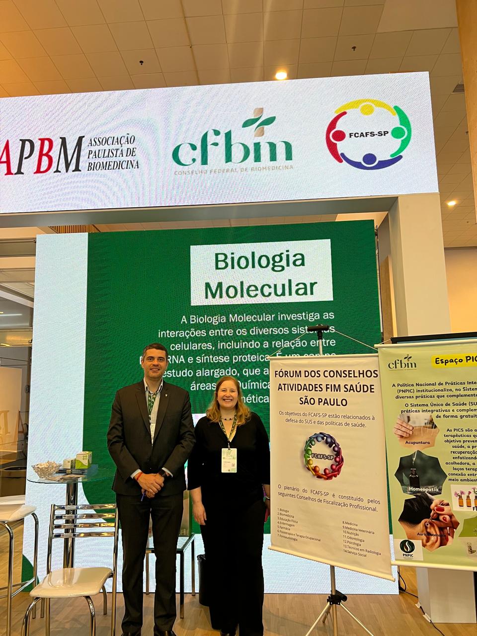 Homem e mulher posando para foto em fachada de evento.