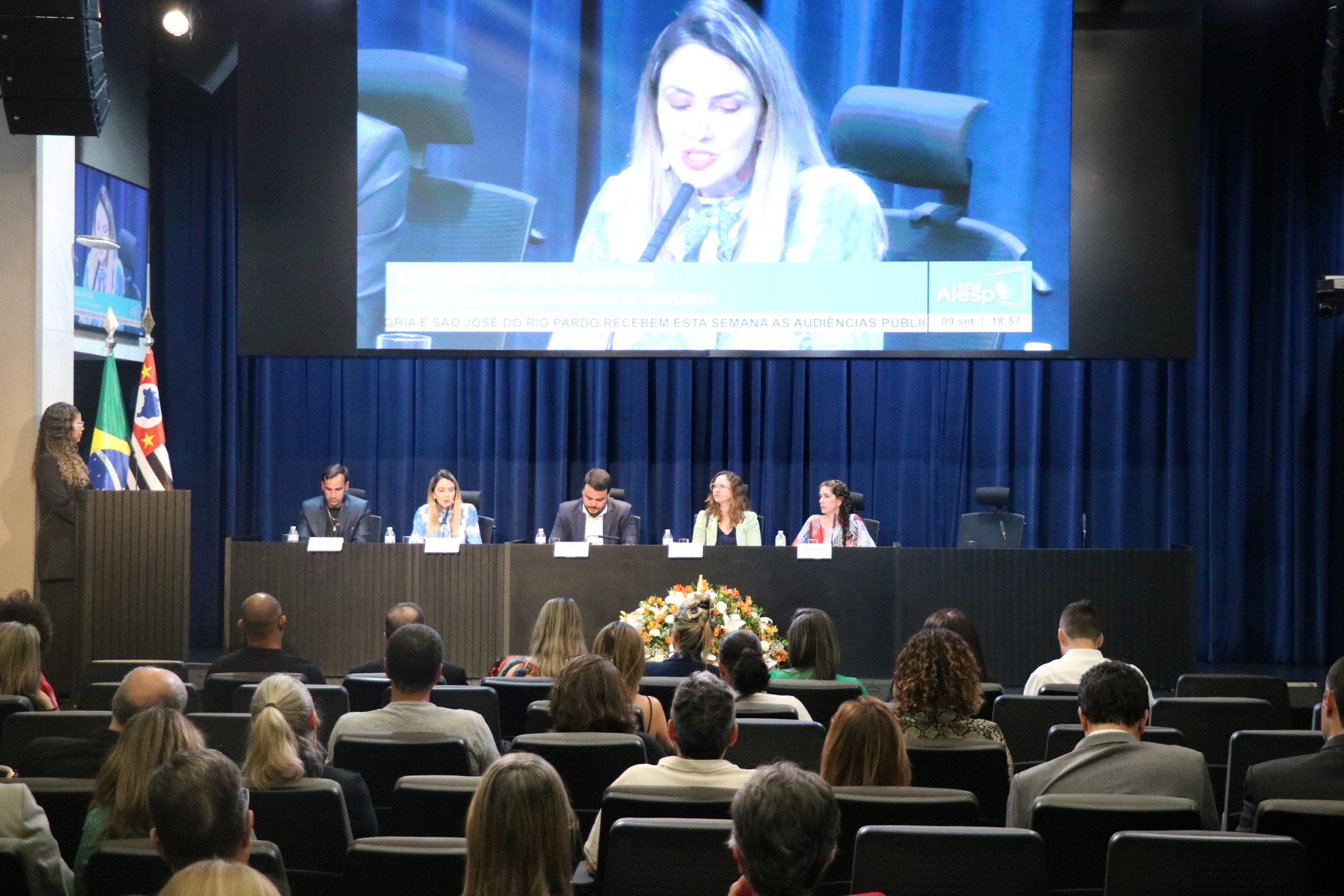 Autoridades sentadas no palco em auditório oficial na Alesp. Da esquerda para a direita: Em pé no púlpito, a coordenadora técnica médica-veterinária do CRMV-SP, Carla Maria Figueiredo de Carvalho e sentados: o secretário-geral do CRMV-SP, Rodrigo Mainardi, a presidente da Comissão de Atenção à Saúde Mental dos Médicos-veterinários e Zootecnistas do CFMV, Ingrid Machado, o secretário de Desenvolvimento Econômico e Trabalho da Prefeitura de São Paulo, Rodrigo Goulart, a presidente do CRMV-SP, Daniela Pontes Chiebao e a vice-presidente do CRMV-SP, Carolina Filippos.