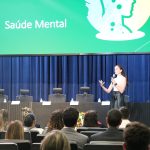 Mulher branca jovem vestindo blusa branca e saia preta longa palestrando em cima de um palco, para uma plateia em auditório na Alesp sobre Saúde Mental. É a psicóloga Bianca Stevanin Gresele.