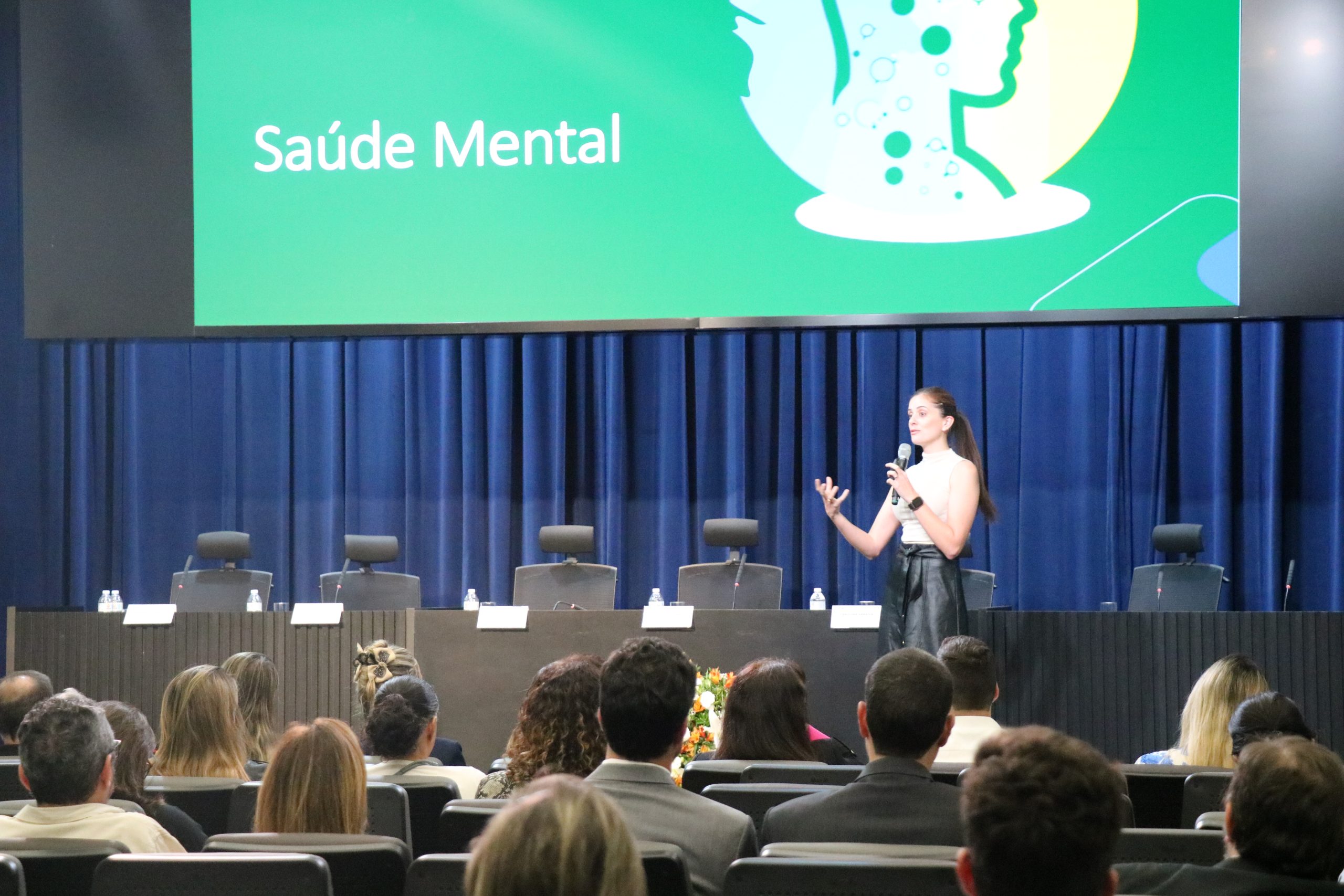 Mulher branca jovem vestindo blusa branca e saia preta longa palestrando em cima de um palco, para uma plateia em auditório na Alesp sobre Saúde Mental. É a psicóloga Bianca Stevanin Gresele.