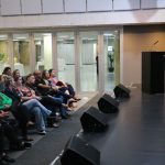 Mulher branca jovem vestindo blusa branca e saia preta longa palestrando em cima de um palco, para uma plateia em auditório na Alesp. É a psicóloga Bianca Stevanin Gresele.