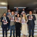 Oito pessoas, duas mulheres e seis homens, reunidas em escadaria segurando papel para foto.