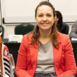 Na foto há uma mulher sorrindo, sentada, de blazer.