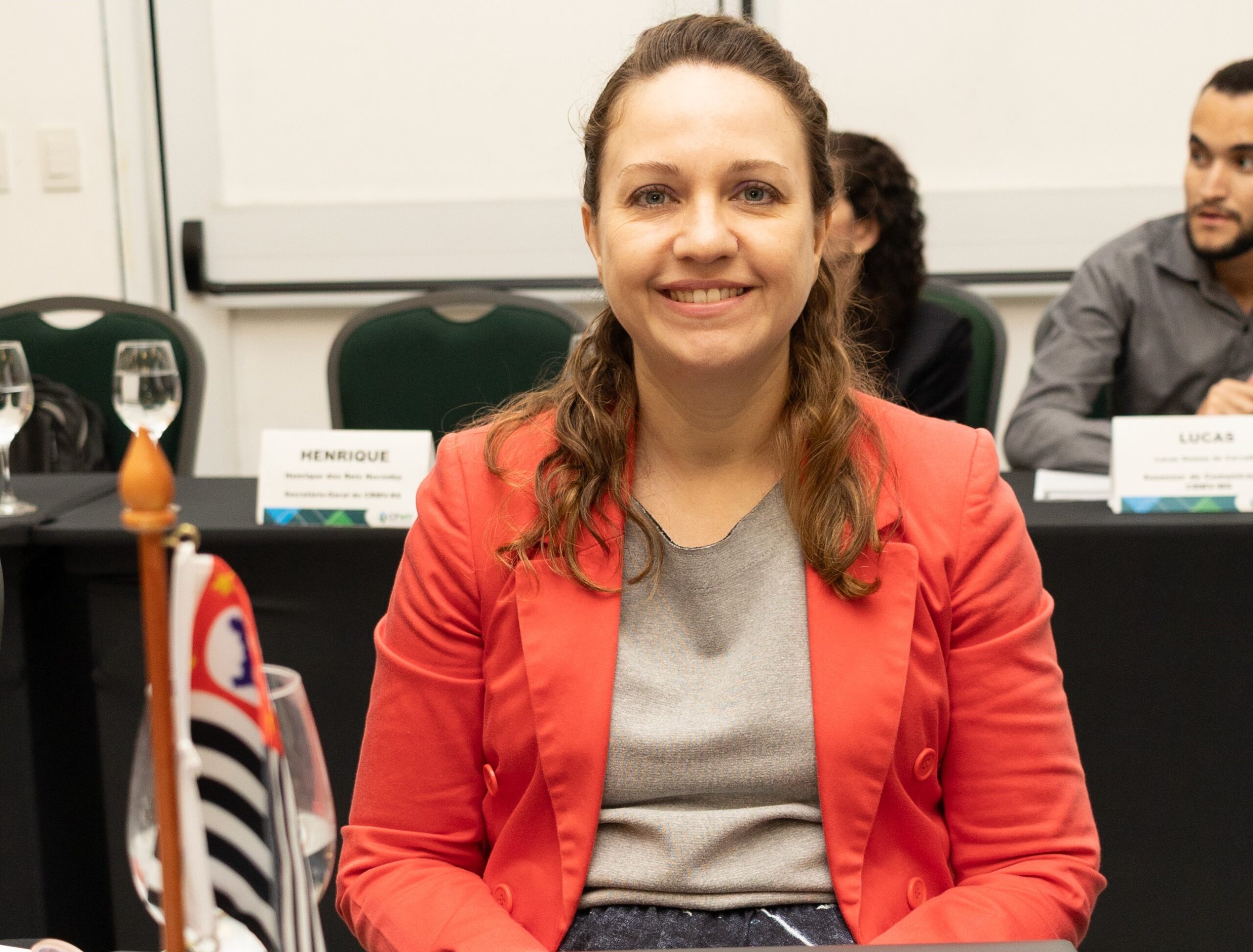 Na foto há uma mulher sorrindo, sentada, de blazer.