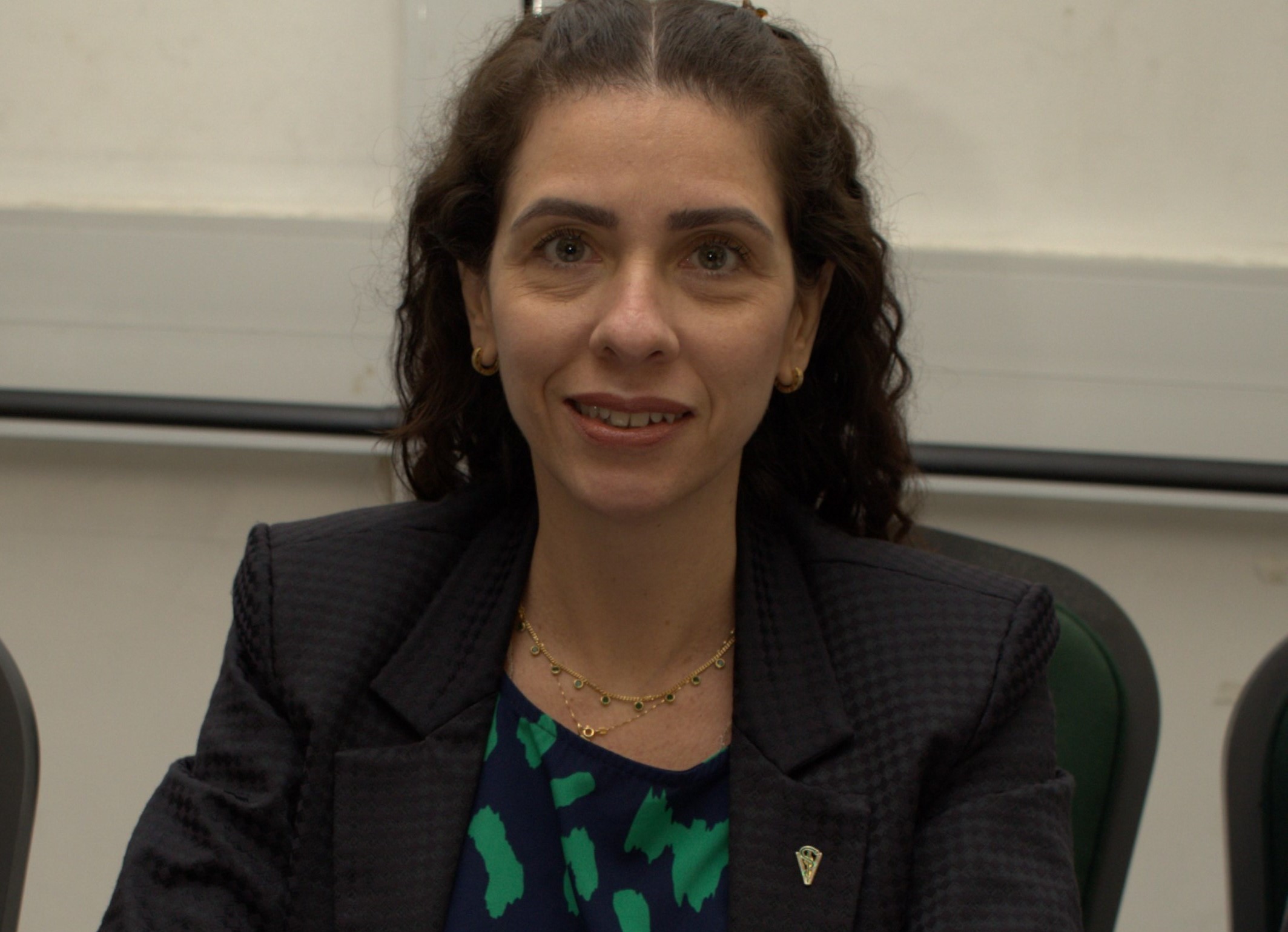 Na foto há uma mulher sentada, sorrindo, de blazer preto.