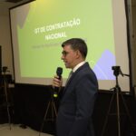 Na foto está um homem de terno falando ao microfone frente ao telão.
