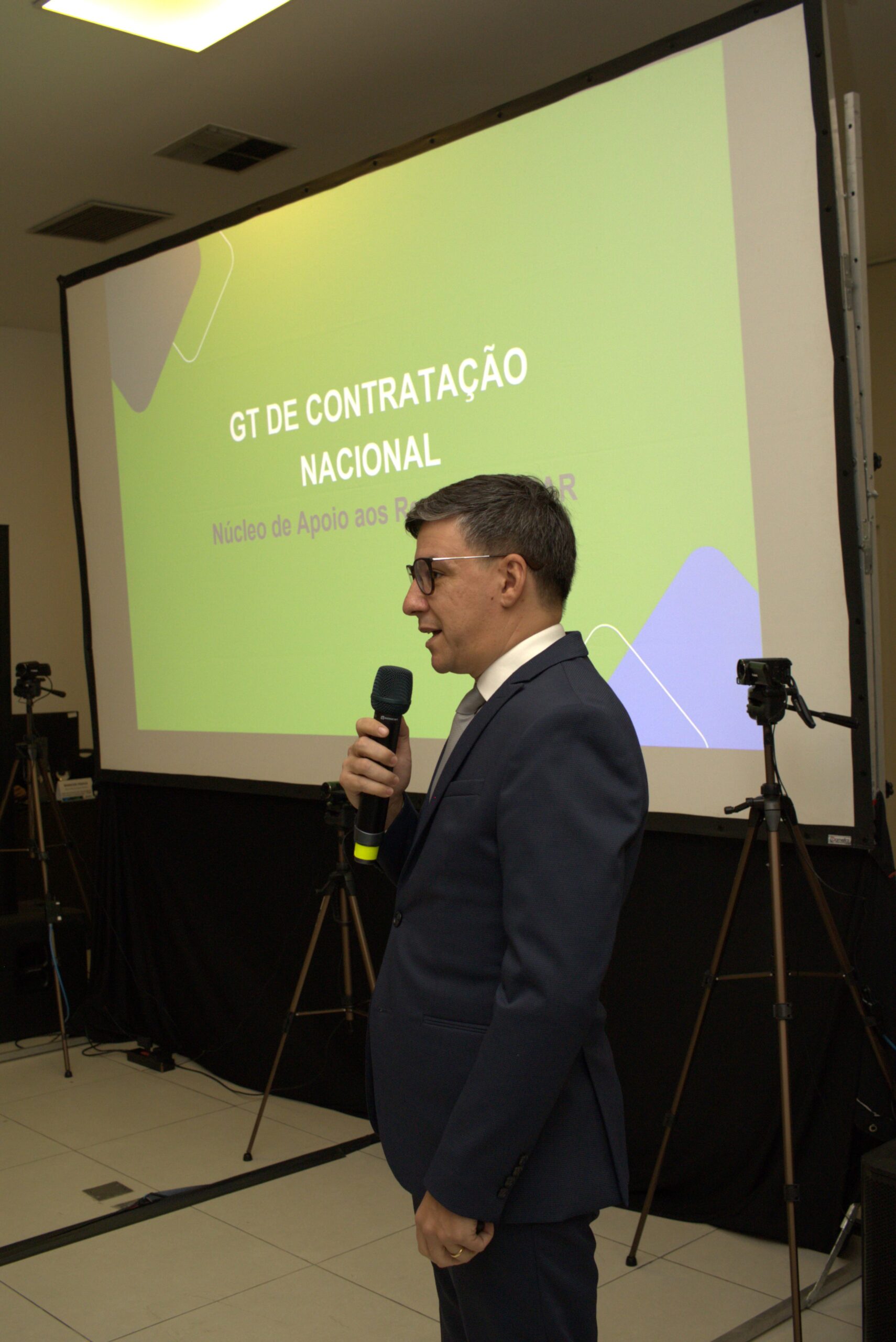 Na foto está um homem de terno falando ao microfone frente ao telão.