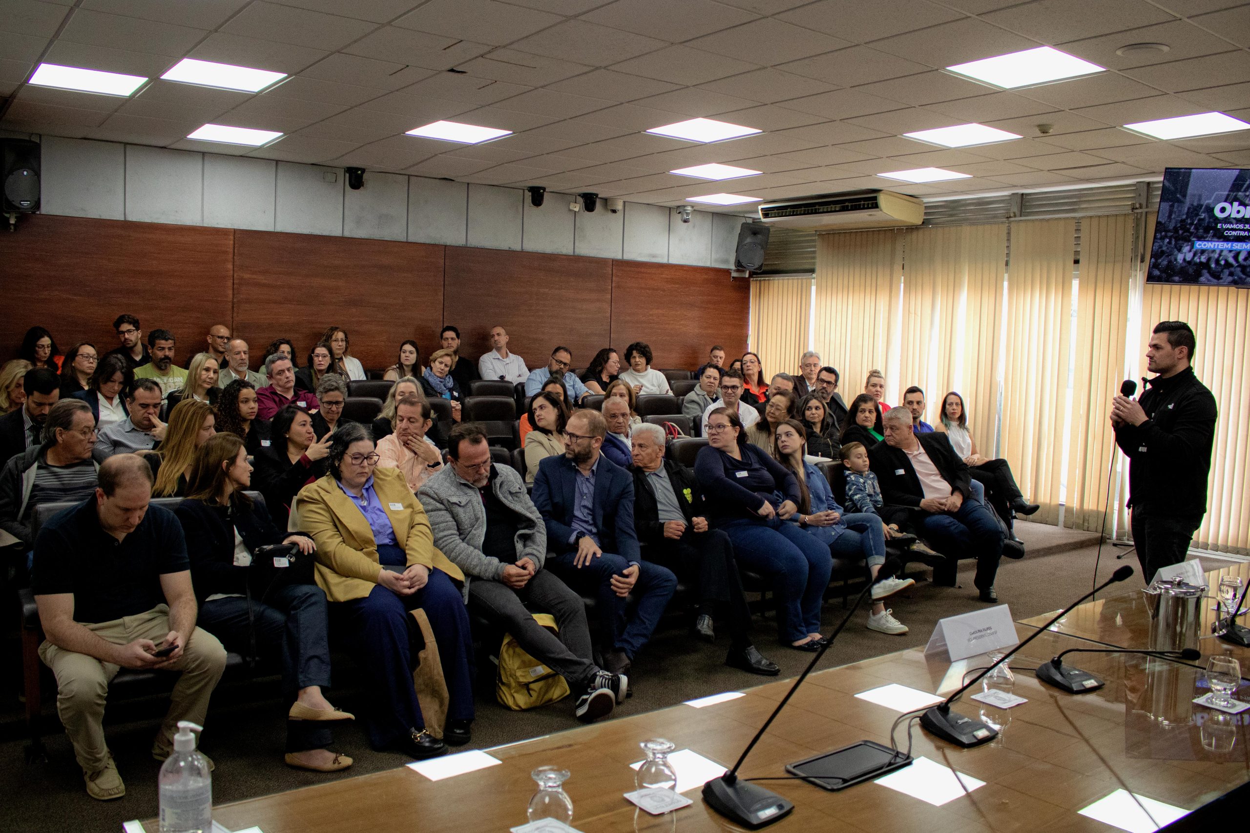 Pessoas sentadas assistindo a uma pessoa falando ao microfone.