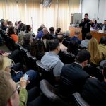 Pessoas sentadas assistindo a uma pessoa falando ao microfone.