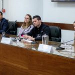 Pessoas sentadas apoiando em uma mesa com microfones.