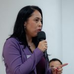 Mulher branca de cabelo preto liso e médio, vestindo blazer roxo palestrando com um microfone na mão.