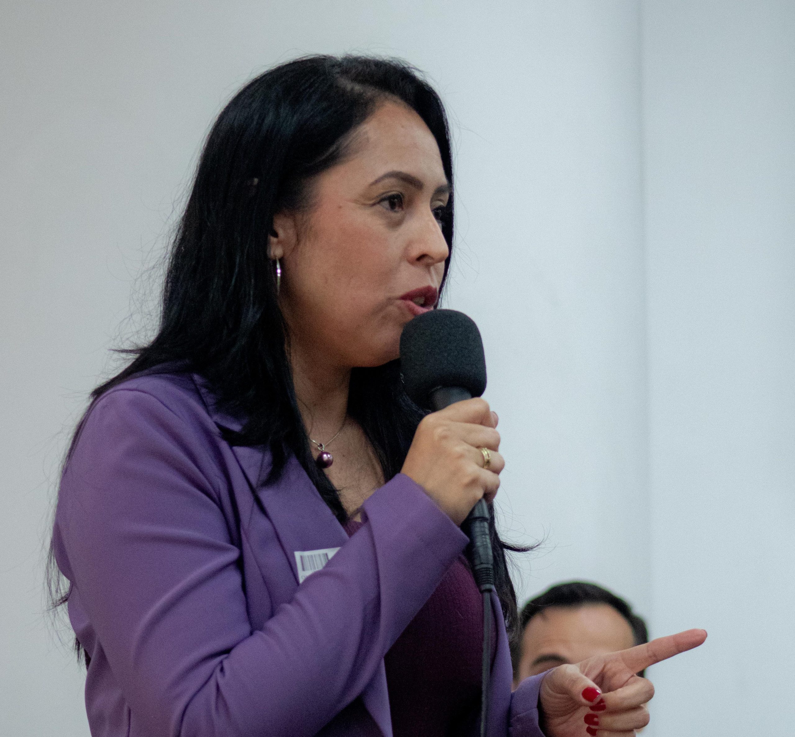 Mulher branca de cabelo preto liso e médio, vestindo blazer roxo palestrando com um microfone na mão.