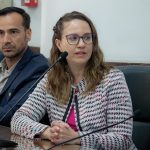 Mulher branca de olhos claros e cabelo loiro escuro usando óculos com armação roxa. Ela veste casaco branco estampado nas cores preto e vermelho e blusa magenta, sentada falando no microfone. É a presidente do CRMV-SP, Daniela Pontes Chiebao.