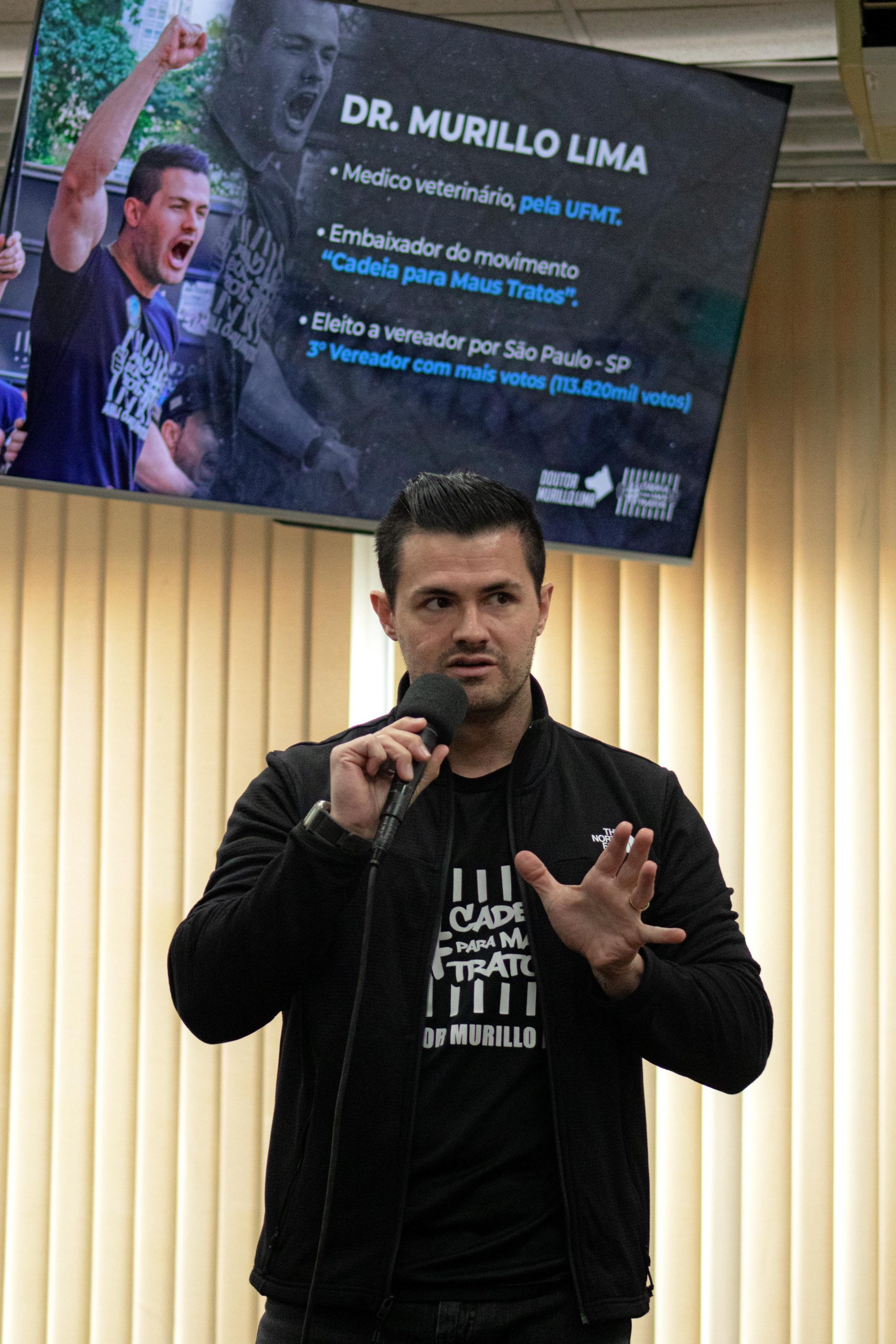Homem branco de olhos pretos e cabelo preto liso curto vestindo blusa de algodão preta com a estampa em letras brancas: "Cadeia para Maus Tratos, Dr. Murillo Lima." e casaco preto, falando no microfone. É o vereador e médico-veterinário por São Paulo, Murillo Lima.