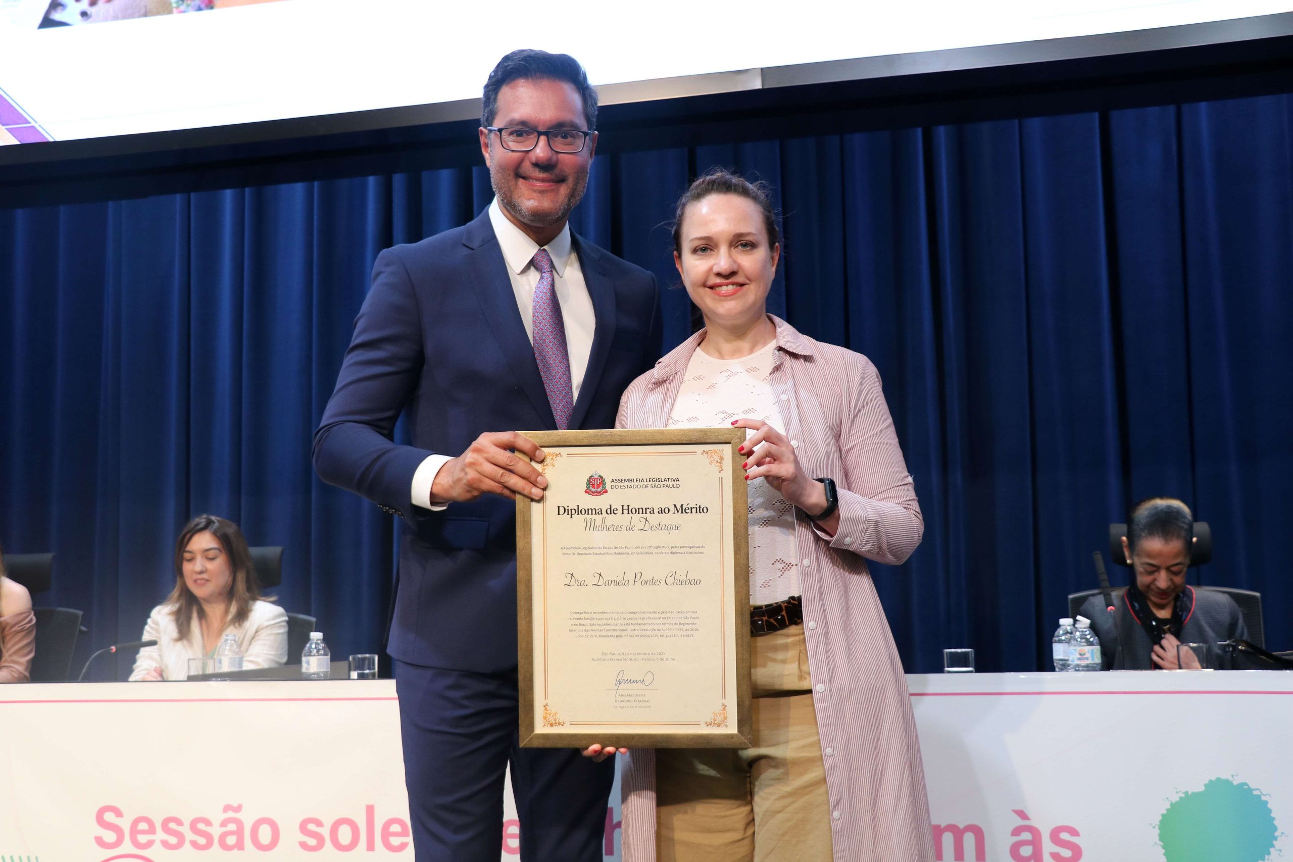Homem e mulher em palco posando para foto segurando placa de homenagem. A mulher é a presidente do CRMV-SP, Daniela Pontes Chiebao.