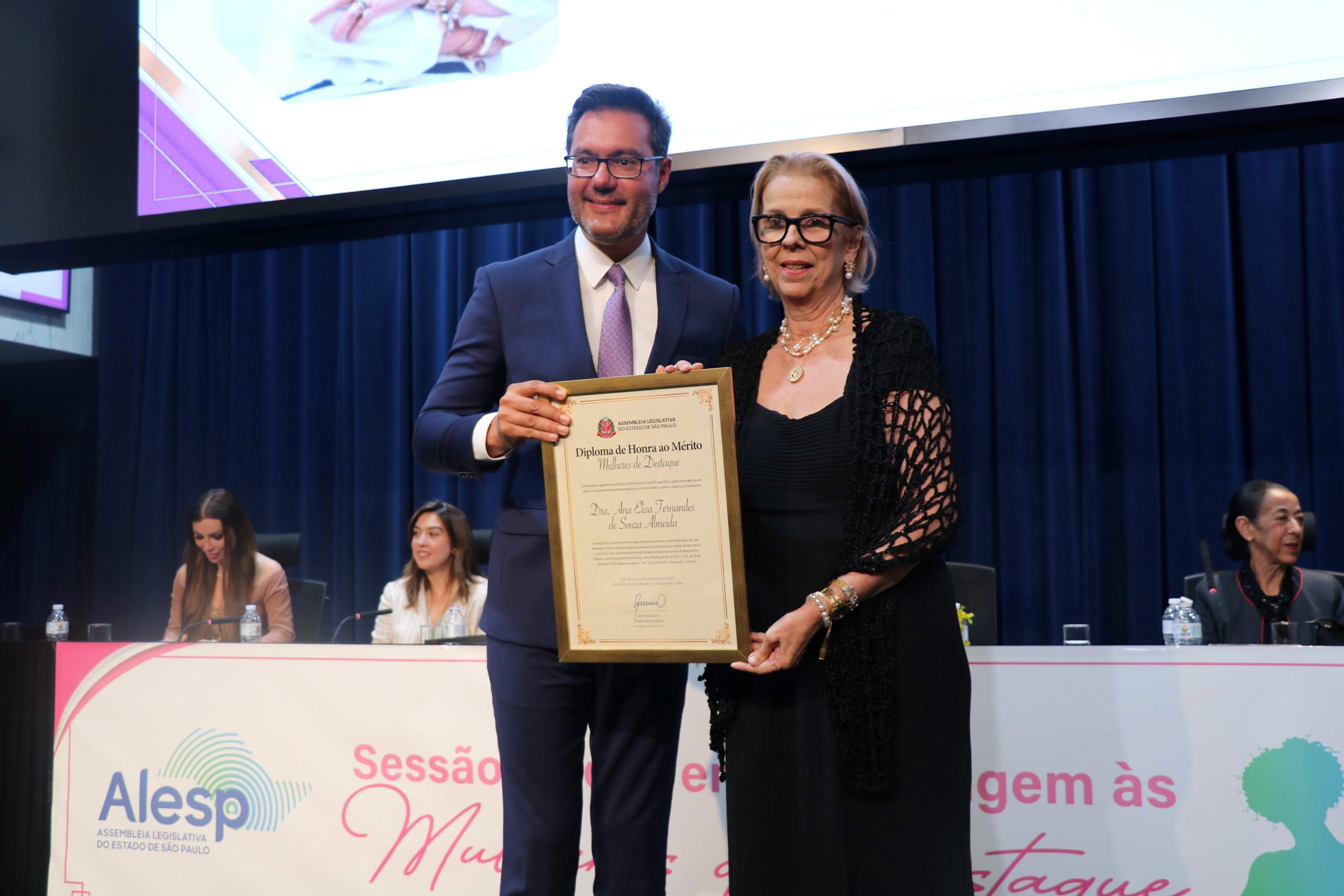 Homem e mulher em palco posando para foto segurando diploma de homenagem. A mulher é a presidente do CFMV, Ana Elisa Fernandes de Souza Almeida.