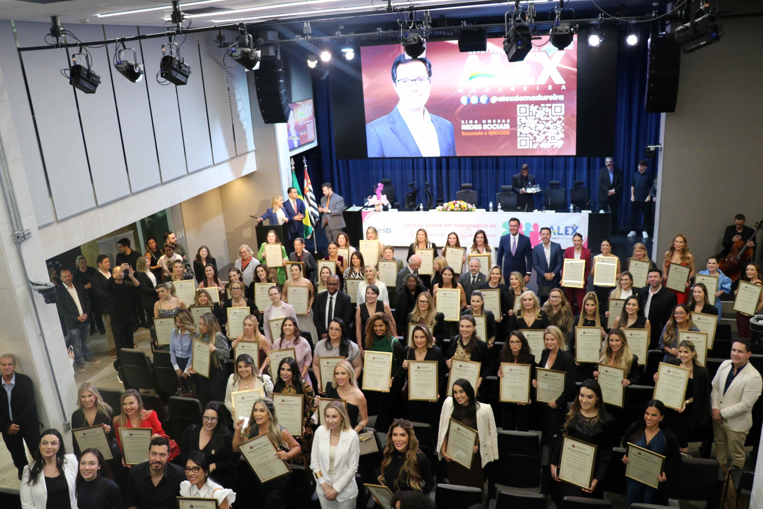 Pessoas reunidas em auditório na Alesp posando para foto segurando diplomas.