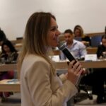 Mulher falando no microfone frente ao público.