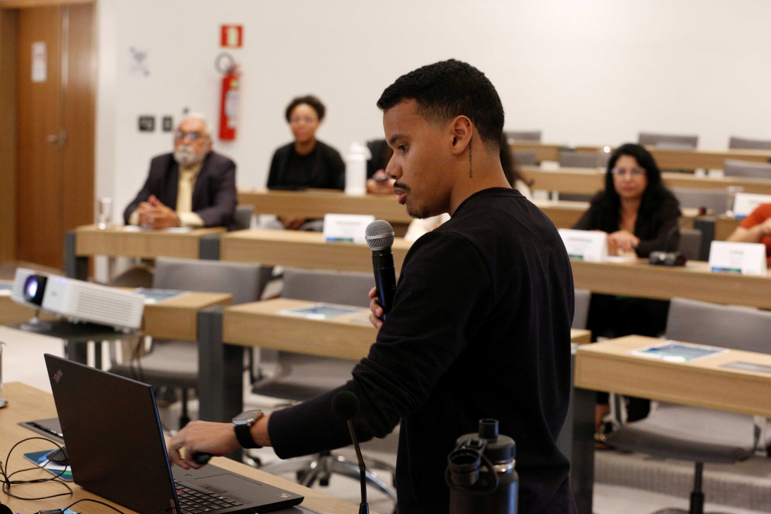 Homem falando no microfone frente ao público.