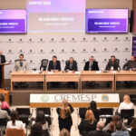 Auditório com mesa contendo sete pessoas ao centro, e outras sentadas participando.