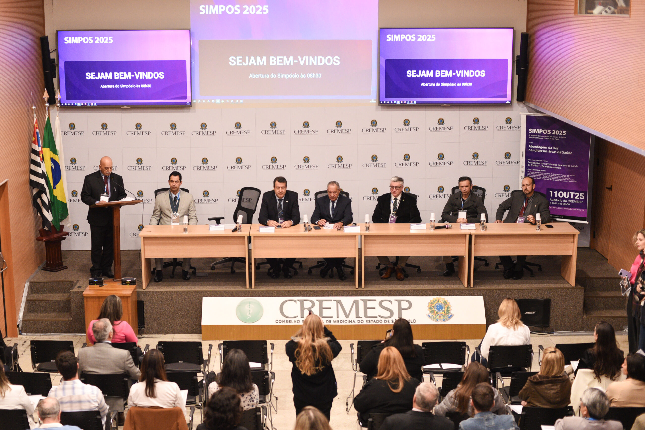 Auditório com mesa contendo sete pessoas ao centro, e outras sentadas participando.
