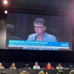 Pessoas sentadas em mesa no palco frente ao telão.