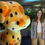 Mulher sorrindo de pé, ao lado de uma escultura grande de gato, em cor laranja.