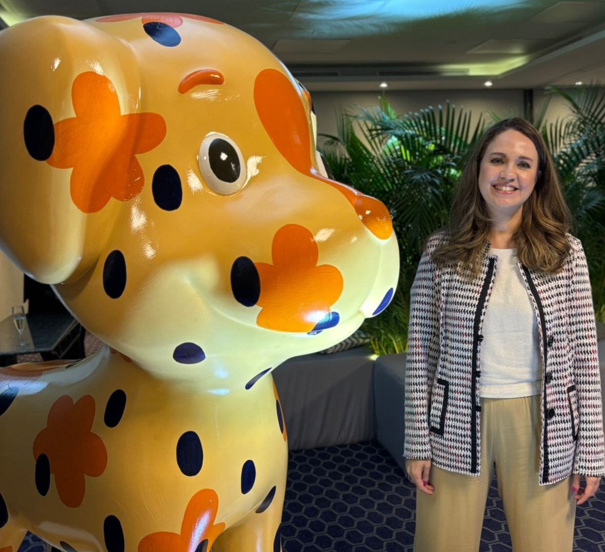 Mulher sorrindo de pé, ao lado de uma escultura grande de gato, em cor laranja.