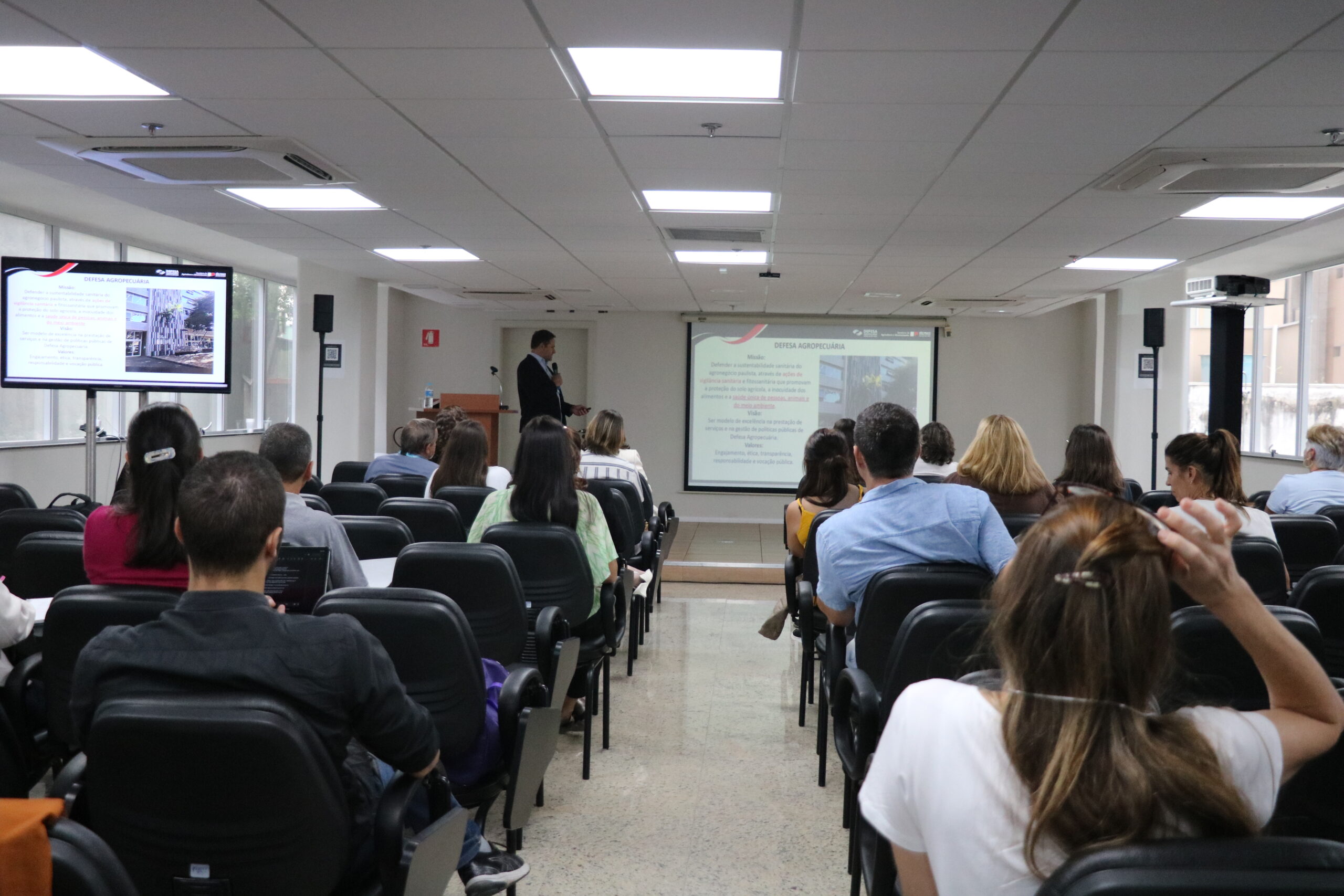 Pessoas sentadas frente ao palco assistindo palestra.