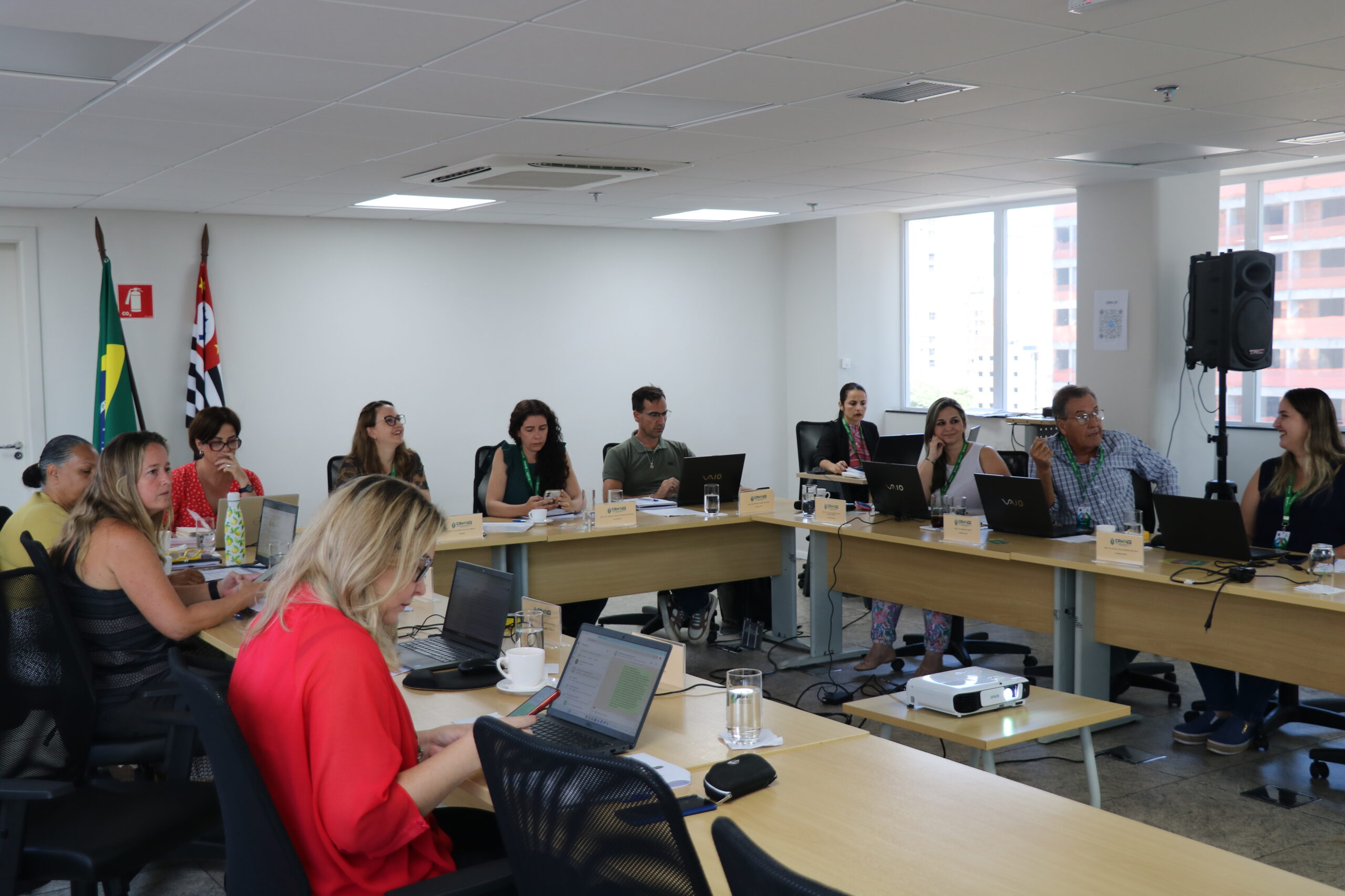 Em uma mesa formato U, homens e mulheres estão sentados e conversando, em reunião.