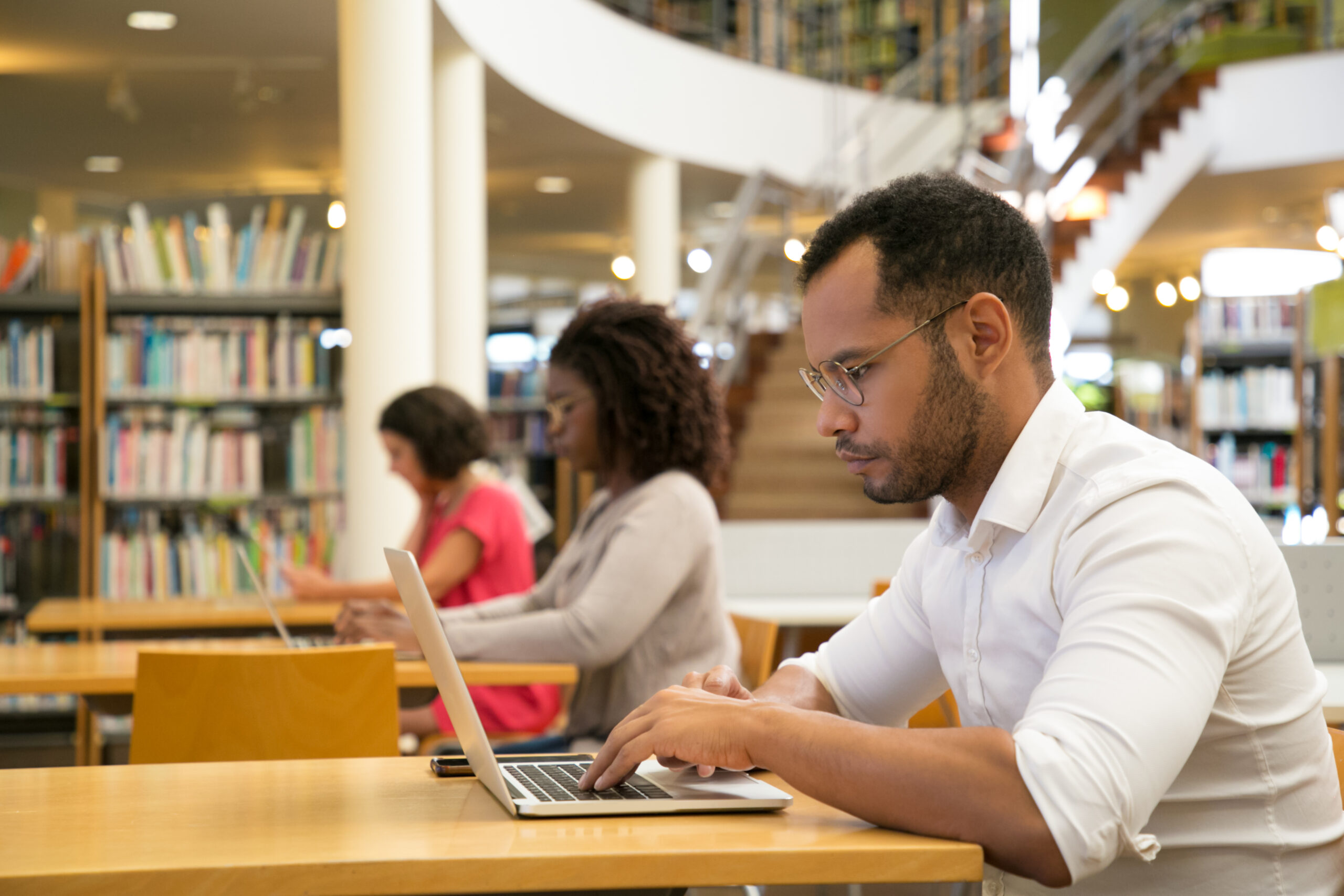 Três pessoas estão sentadas a frente de computadores no ambiente de estudos de uma biblioteca. Em destaque está um homem jovem com cabelos curtos e barba bem aparada utilizando óculos. Todos são vistos de perfil.