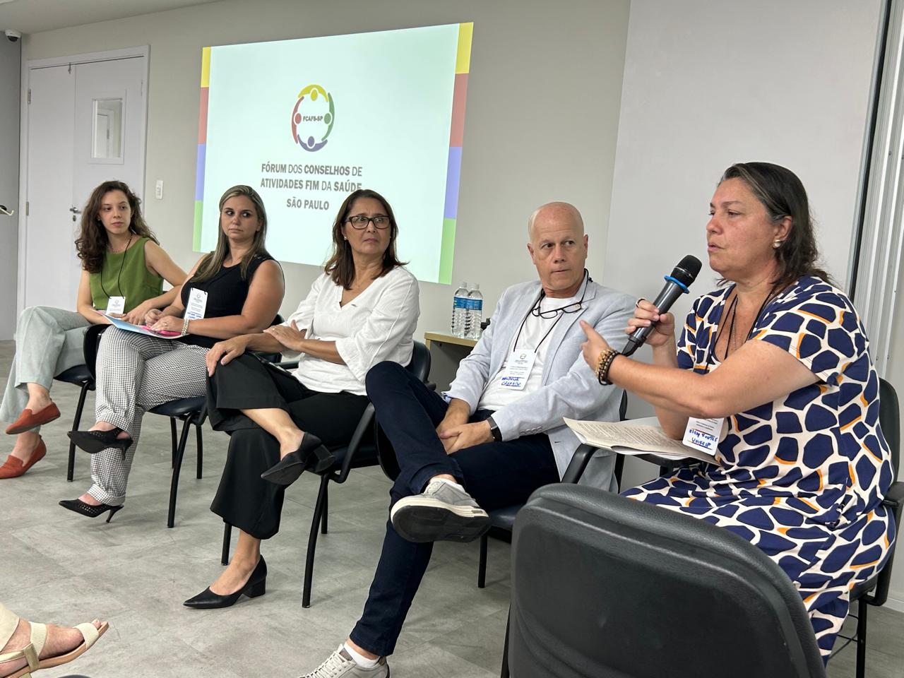 Mulher sentada com o microfone na mão junto de outras quatro pessoas em palestra durante evento do Fcafs-SP