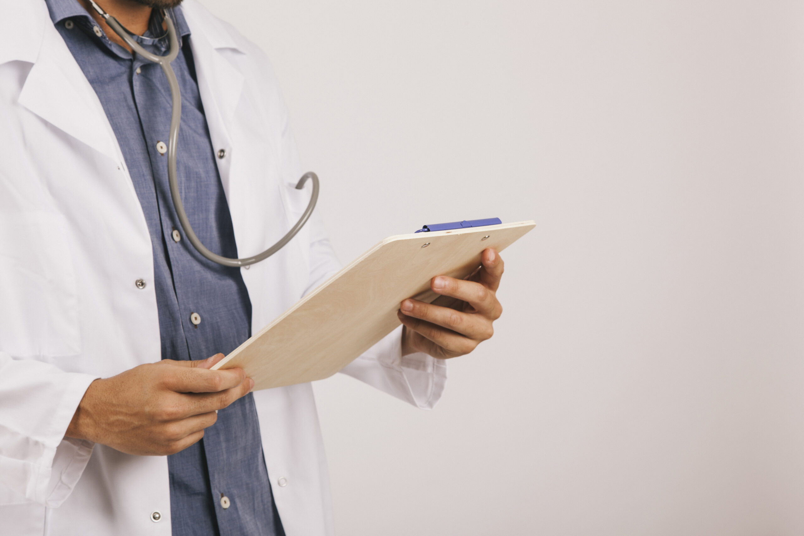 Médico-veterinário está de camisa azul e jaleco branco, estetoscópio pendurado no pescoço e segura uma prancheta com as duas mãos.
