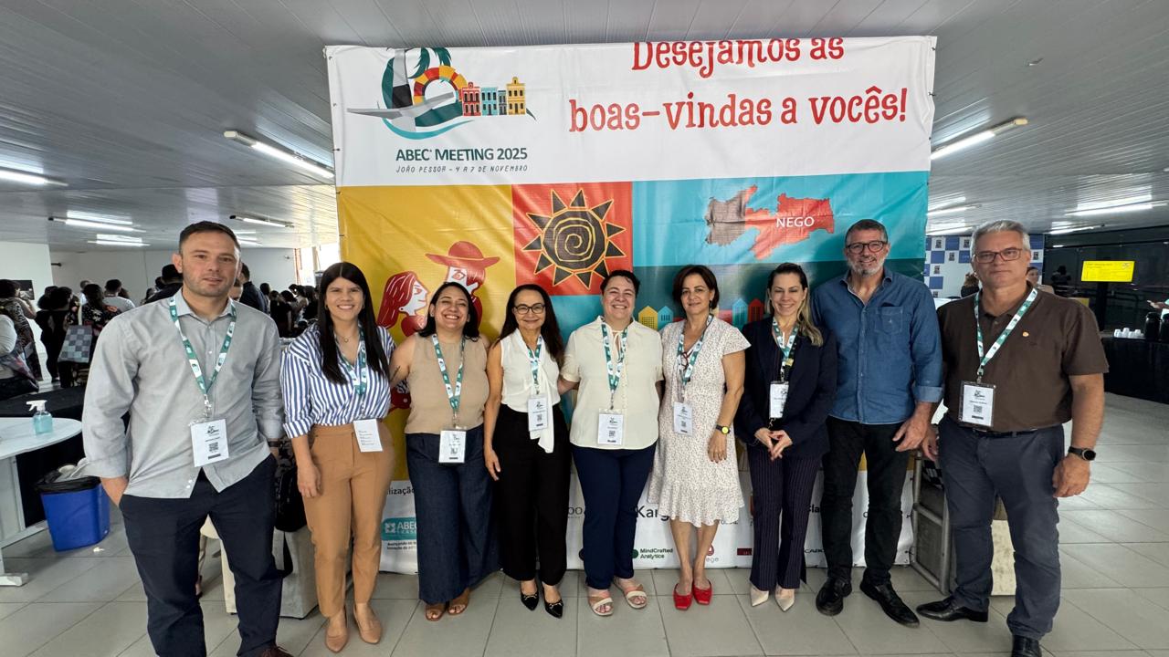 Integrantes das Comissões Editoriais do CFMV e do CRMV-SP posam de pé para foto, em frente ao painel de entrada do Abec Meeting. O painel é colorido com desenhos que lembram xilografias nordestinas. 