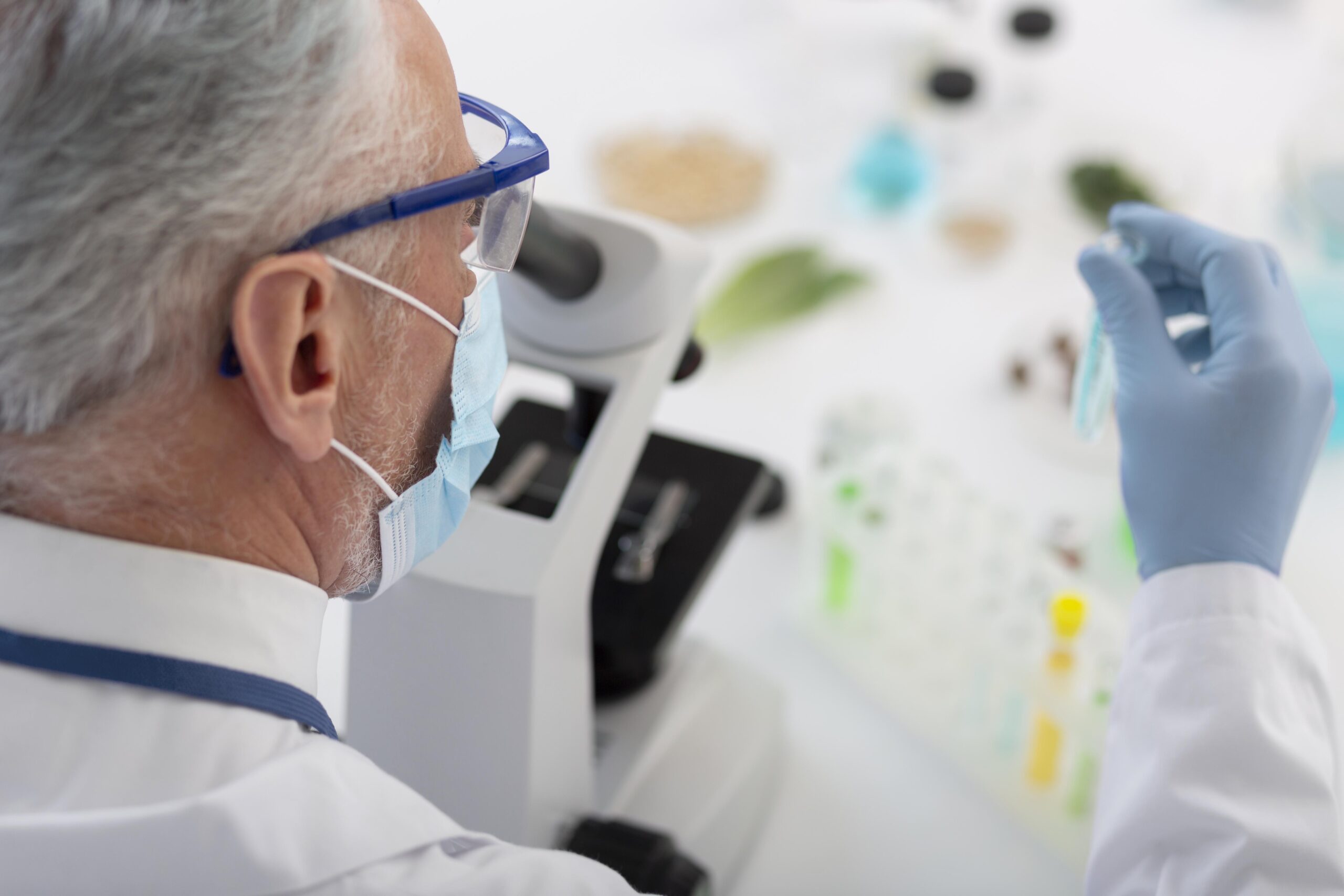 Homem grisalho analisa amostras de laboratório em um microscópio. Ele usa máscara, luvas e óculos de proteção. 