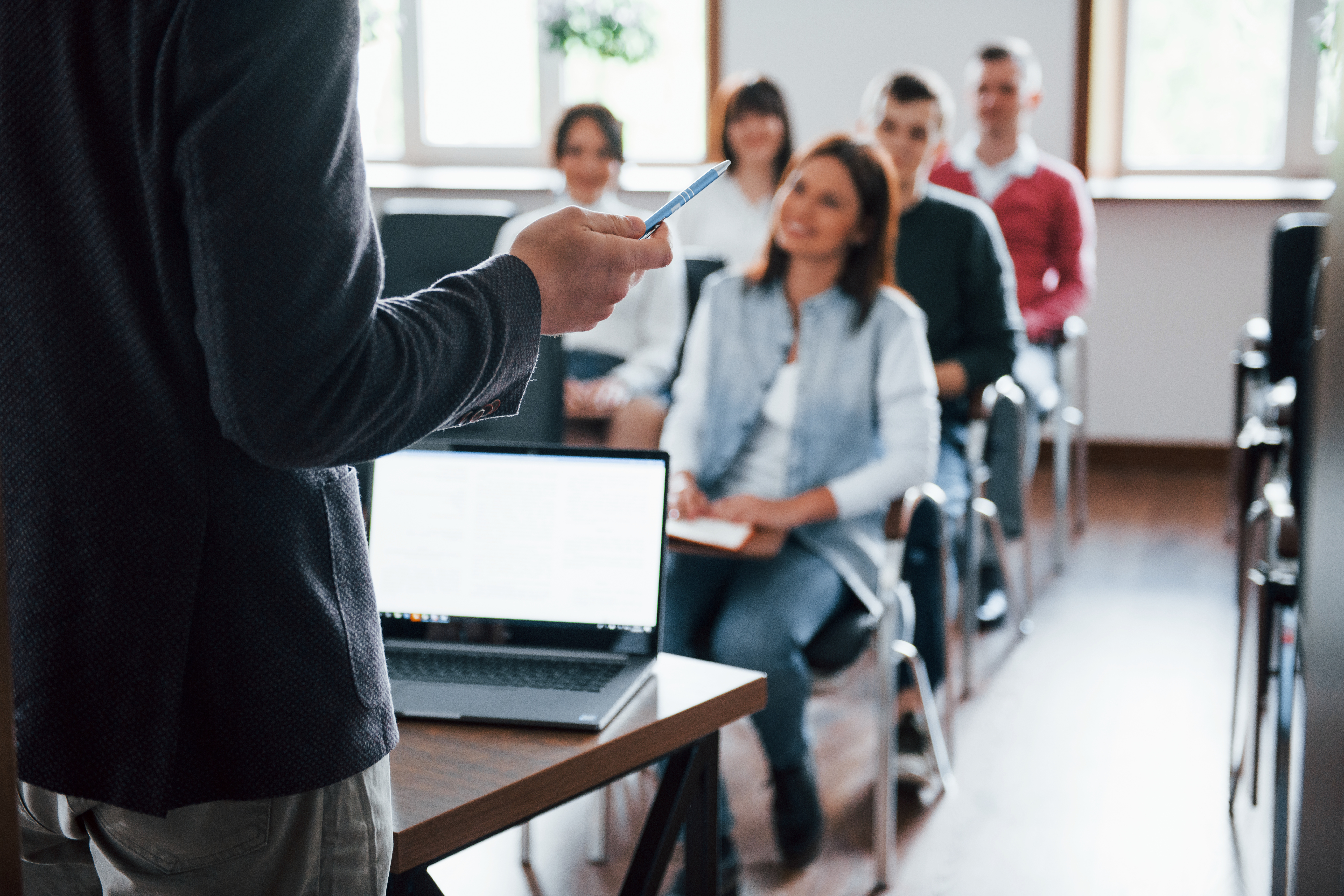 Imagem de uma sala de aula. O professor está a frente, de costas para a foto, e os alunos aparecem sentados.