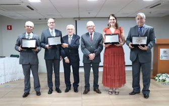 Seis pessoas em pé, sendo cinco homens e uma mulher. Eles estão lado a lado, quatro deles estão segurando placas de homenagem