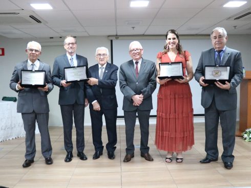 Seis pessoas em pé, sendo cinco homens e uma mulher. Eles estão lado a lado, quatro deles estão segurando placas de homenagem