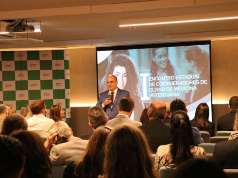 Na imagem há uma auditório de eventos. O vice-presidente do Conselho está a frente de pé e com um microfone em mão, falando para uma plateia sentada. Atrás dele há um painel quadriculado verde e branco com a logomarca do CRMV-SP e um telão com o nome do evento: I Encontro Estadual de Coordenadores de Curso de Medicina Veterinária