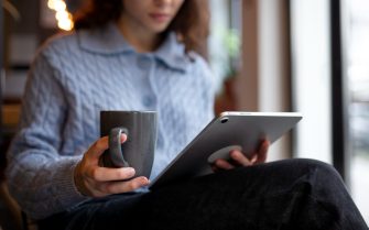Mulher branca lendo revista científica em um tablet cinza, com uma caneca cinza na mão esquerda. Ela veste blusa social azul de mangas longas e botões ao meio e calça social preta.
