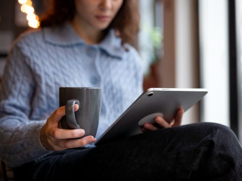 Mulher branca lendo revista científica em um tablet cinza, com uma caneca cinza na mão esquerda. Ela veste blusa social azul de mangas longas e botões ao meio e calça social preta.