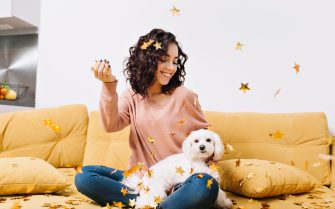 Happy weekends, true positive emotions of young joyful woman with cut curly hair having fun with little dog in falling golden tinsels on couch in modern apartment