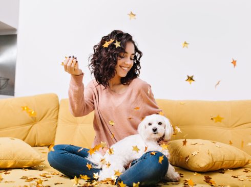 Happy weekends, true positive emotions of young joyful woman with cut curly hair having fun with little dog in falling golden tinsels on couch in modern apartment