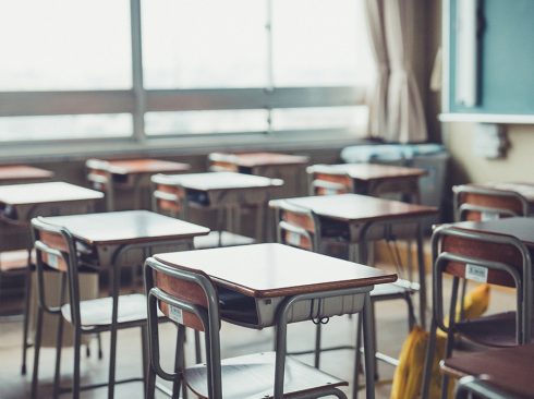 Sala de aula com várias carteiras escolares e uma ampla janela ao fundo. Há também uma lousa verde.