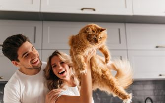 Casal jovem e branco, vestindo blusas brancas de mangas curtas e gola redonda segura gato laranja adulto de pelo espesso. Eles estão em uma cozinha.