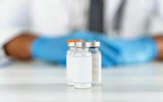 3 Frascos de remédio com uma pessoa ao fundo usando uniforme e luvas brancas.