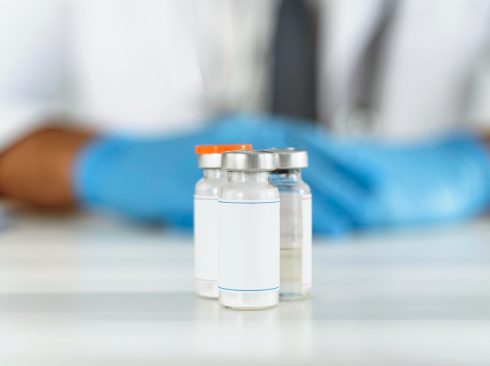 3 Frascos de remédio com uma pessoa ao fundo usando uniforme e luvas brancas.