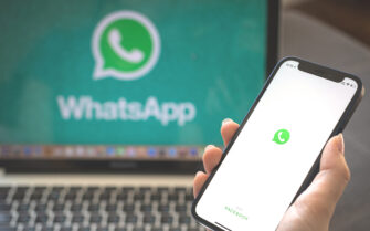 Pessoa com um celular na mão, em que aparece o ícone do WhatsApp. Ao fundo, um computador de mesa, com o símbolo da rede social também aparecendo na tela.